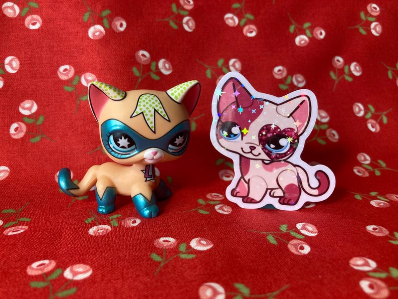 May include: Two toy cats, one with a superhero mask and the other with a pink heart on its eye, are on a red background with a floral pattern.