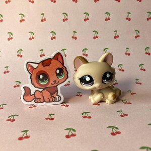 K&ouml;nnte beinhalten: Zwei kleine Plastik-Spielzeugkatzen. Eine ist rot und wei&szlig; mit gr&uuml;nen Augen und die andere ist beige mit violetten Augen. Beide sitzen auf einem rosa Hintergrund mit einem Kirschmuster.