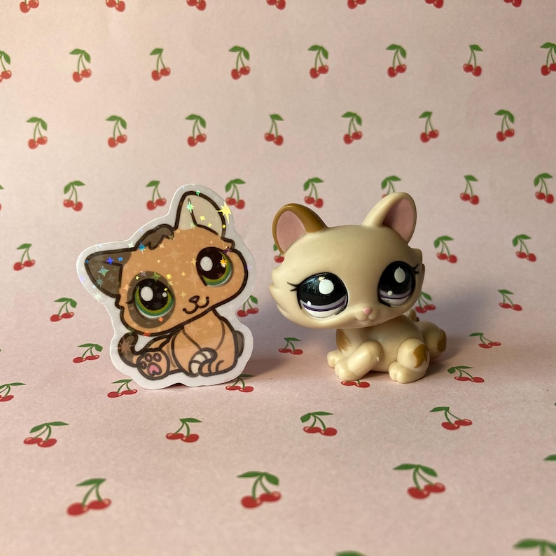 K&ouml;nnte beinhalten: Zwei kleine Plastik-Spielzeugkatzen. Eine ist ein brauner und wei&szlig;er Comic-Katzenaufkleber. Die andere ist eine beige Katze mit braunen Ohren und lila Augen.