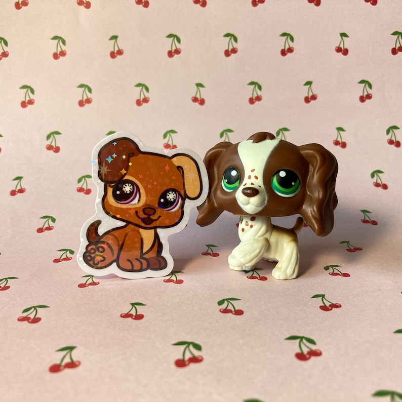 K&ouml;nnte beinhalten: Zwei kleine braune und wei&szlig;e Spielzeug-Hunde. Ein Hund ist ein Aufkleber mit glitzernden Augen und der andere ist eine 3D-Figur.