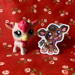 May include: A pink and white toy animal with a brown and white toy animal sticker. The sticker has a brown cow with white spots and pink hearts for eyes. The toy animals are on a red background with white flowers.