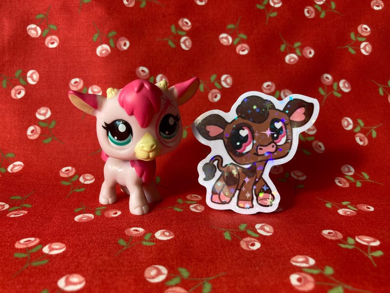 May include: A pink and white toy animal with a brown and white toy animal sticker. The sticker has a brown cow with white spots and pink hearts for eyes. The toy animals are on a red background with white flowers.