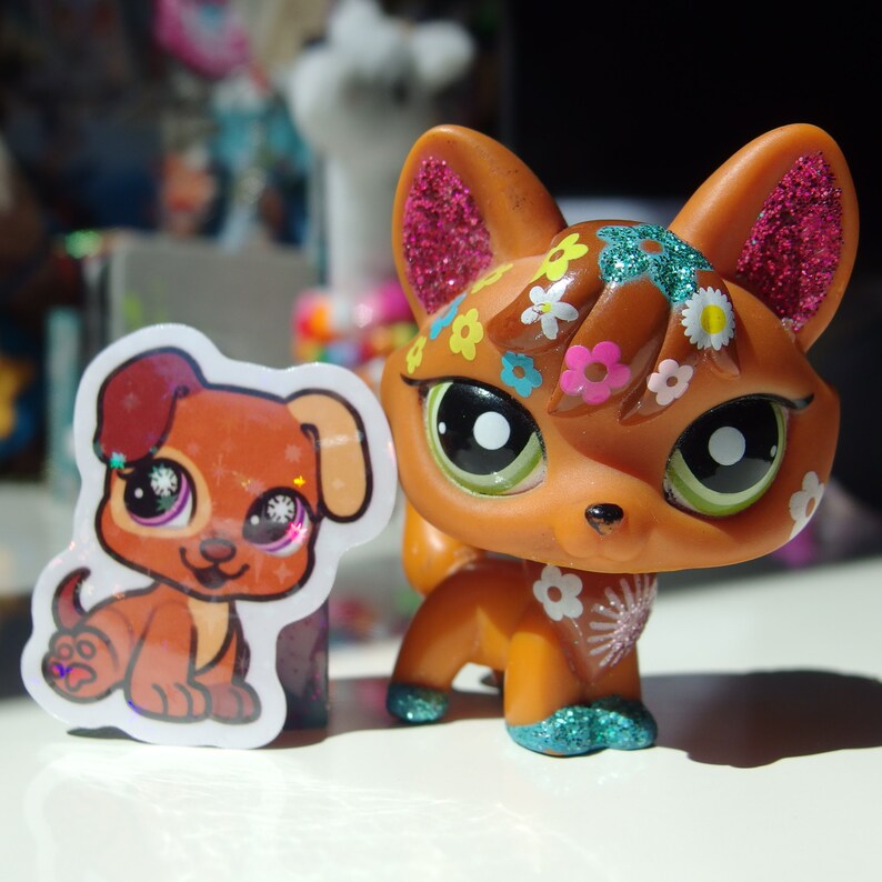 May include: A brown fox figurine with glitter and flower accents. The fox has green eyes and a pink nose. The figurine is standing on a white surface.