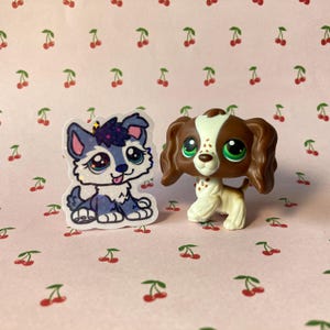 K&ouml;nnte beinhalten: Zwei kleine Plastikspielzeug-Hunde. Einer ist ein blauer und wei&szlig;er Husky mit einem lila Stern auf dem Kopf. Der andere ist ein brauner und wei&szlig;er Spaniel mit gr&uuml;nen Augen.