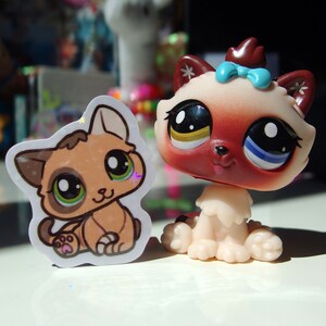 May include: Two plush toy cats. One is a brown and white cat with green eyes and a pink paw print. The other is a red and white cat with blue and yellow eyes and a blue bow.