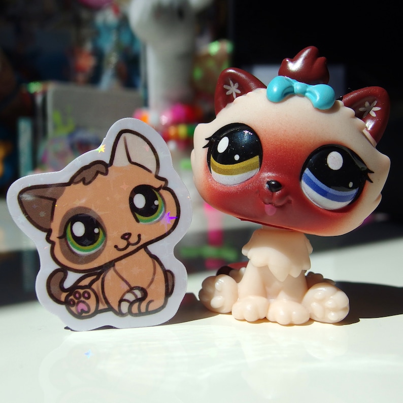 May include: Two plush toy cats. One is a brown and white cat with green eyes and a pink paw print. The other is a red and white cat with blue and yellow eyes and a blue bow.
