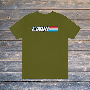 Peut inclure: T-shirt vert olive avec le mot "LINUX" en blanc, souligné de noir, au-dessus d'un motif à rayures rouges, blanches et bleues. Le t-shirt est en matière souple et est présenté sur une surface en bois.