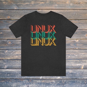 May include: Dark grey t-shirt featuring the word "LINUX" stacked three times in red, teal, and gold. The text has a retro, layered design. The shirt is displayed on a wooden surface.