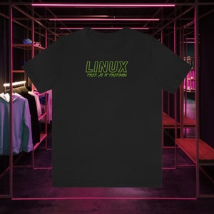 Puede incluir: Camiseta negra con la palabra "LINUX" en verde neón, sobre la frase "FREE AS IN FREEDOM". La camiseta se muestra en un entorno comercial con percheros y luces de neón.