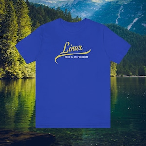 May include: A royal blue t-shirt featuring the word "Linux" in a yellow script font, underlined with a yellow curve. Beneath, the text reads "Free as in Freedom". The backdrop shows a lake and mountains, creating a scenic setting.