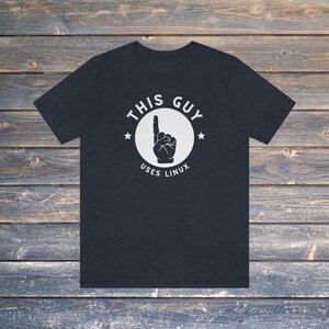 May include: Dark navy blue t-shirt with a white graphic. The graphic shows a hand pointing upwards inside a circle, with the text "THIS GUY USES LINUX". Two stars are on either side of the circle. The shirt is made of a soft material.