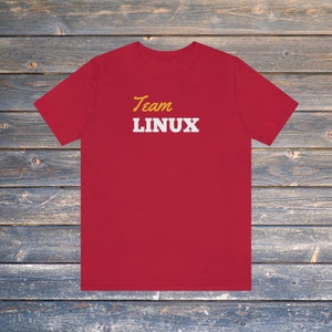 Pode incluir: Uma t-shirt vermelha com a frase "Team LINUX" impressa. A palavra "Team" está em itálico amarelo, e "LINUX" em negrito branco. A t-shirt é exibida sobre uma superfície de madeira.