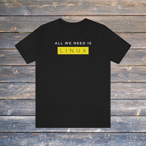 May include: Black t-shirt featuring the text "ALL WE NEED IS LINUX" in white above a yellow rectangle with the word "LINUX" in white. The shirt is shown on a wooden background.