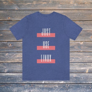 May include: A navy blue t-shirt featuring the words "JUST USE LINUX" in white, each word separated by a red bar. The tee is set against a wooden backdrop.