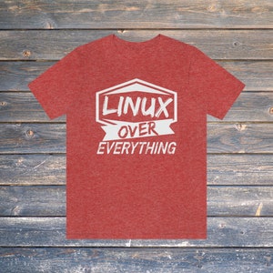May include: Red t-shirt featuring the white text "LINUX OVER EVERYTHING". The text is within a white hexagon and banner. The shirt is made of a soft material and is displayed on a wooden background.