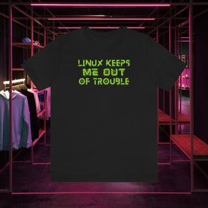 Może przedstawiać: Czarna koszulka z zielonym napisem "LINUX KEEPS ME OUT OF TROUBLE" w pikselowanej czcionce. Koszulka jest prezentowana w otoczeniu sklepowym, co sugeruje, że jest to produkt do sprzedaży. Projekt jest prawdopodobnie skierowany do entuzjastów komputerów lub Linuksa.