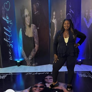 May include: A woman stands in front of a backdrop featuring large portraits. The backdrop includes the text "we still do" and "Jye". A floor graphic displays the name "Janique" and the date "Est. August 19. 2".