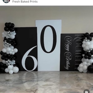 May include: A birthday party decoration set featuring large black and white number cutouts for the number 60. The backdrop includes balloon arrangements in black, white, and silver. The words "Happy Birthday" are also visible.