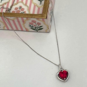 May include: A silver necklace with a heart-shaped pendant. The pendant features a vibrant red gemstone set within a silver frame. The necklace is displayed on a white surface, with a decorative box in the background.