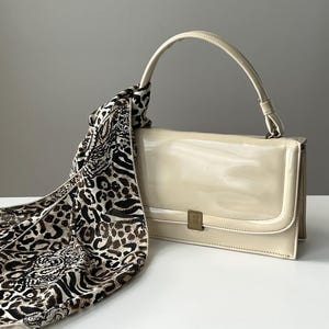 May include: Cream-colored handbag with a top handle and a gold-tone clasp. A patterned scarf with animal print in shades of brown and black is draped over the bag. The bag has a glossy finish.