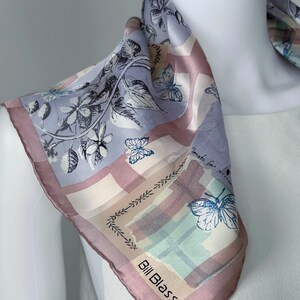 May include: A square silk scarf with a butterfly and floral design. The scarf features a light purple, pink, and cream color palette. The brand name "Bill Blass" is printed on the corner of the scarf.