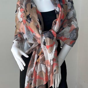 May include: A lightweight scarf featuring a multi-colored abstract pattern in shades of coral, tan, gray, and white. The fabric has a subtle grid texture and is finished with small, dangling tan tassels along the bottom edges of the scarf.