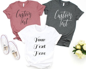 May include: Three t-shirts with customizable text. One is pink, one is gray, and one is white. The pink and gray shirts say "Custom Text" in white script. The white shirt says "Your Text Here" in black script.