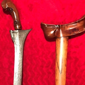 以下が含まれることがあります： 木製の鞘が付いた伝統的なインドネシアのクリス短剣。刃は銀色で、湾曲した柄は濃い木で作られています。鞘は薄い茶色の木で、濃い茶色の筋が入っています。