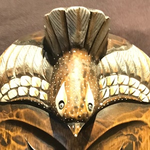May include: A wooden mask with a bird design. The bird has white and brown feathers and a beak. The mask is painted with brown and white paint.