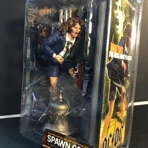 Può includere: Action figure di Angus Young degli AC/DC, confezionata in una custodia di plastica trasparente. La figura indossa un completo blu e pantaloncini, con camicia bianca e cravatta. La confezione include il logo della band e il testo "For Those About to Rock".