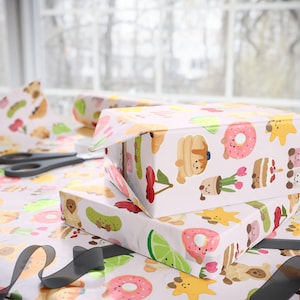 Peut inclure: Deux boîtes cadeaux emballées dans du papier rose clair avec un motif répété d'aliments de dessins animés comme des beignets, des crêpes et des cerises. Les boîtes sont empilées les unes sur les autres. Ciseaux et ruban noir visibles.