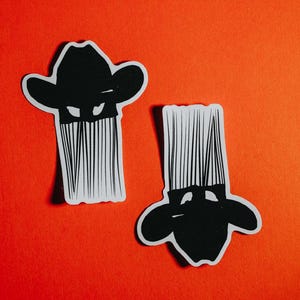 K&ouml;nnte beinhalten: Zwei schwarz-wei&szlig;e Aufkleber einer Person mit Cowboyhut und langem Bart. Die Aufkleber befinden sich auf einem roten Hintergrund.