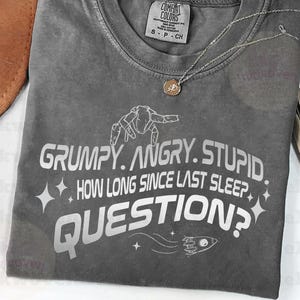 May include: A charcoal gray crew neck t-shirt featuring white graphic text that reads GRUMPY. ANGRY. STUPID. HOW LONG SINCE LAST SLEEP QUESTION? along with a small rocket ship icon and star accents. The shirt has a Comfort Colors brand label.