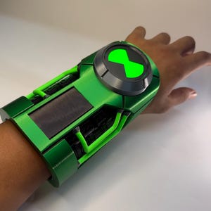 May include: A metallic green wrist-worn device featuring a circular dial with a bright neon green hourglass symbol at the centre. The accessory includes black rectangular panels and green structural piping, designed to fit around the forearm.
