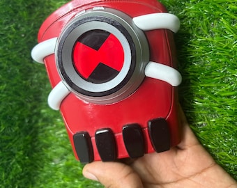 May include: A red and silver Omnitrix toy with a black and red symbol in the center. The toy has white accents and black rectangular details. The toy is a replica of the device from the animated series.