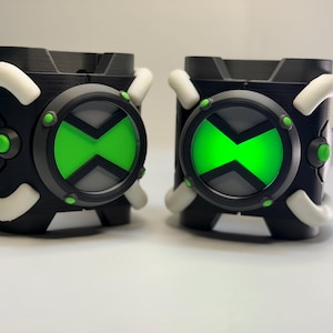 May include: Two black wrist-worn devices featuring a circular centre with a bright green hourglass symbol, surrounded by a dark grey ring with small green accents. Each device has four white curved bars extending from the sides of the main body.