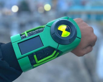 May include: A green and black toy watch with a yellow and black symbol on the face. The watch has a digital display and green tubing details. The watch is worn on a wrist.