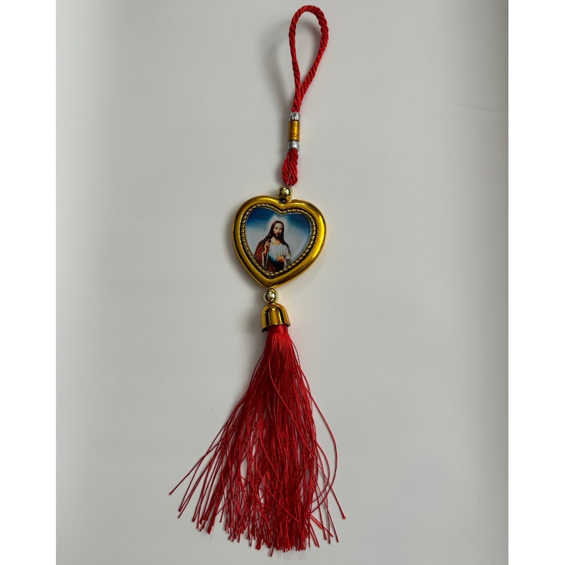 May include: A red tassel hanging from a gold heart-shaped charm with a picture of Jesus inside. The charm is attached to a red string.