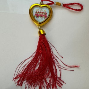 May include: A gold heart-shaped charm with a red tassel. The charm features a white background with a colorful image of five people in red and yellow clothing.