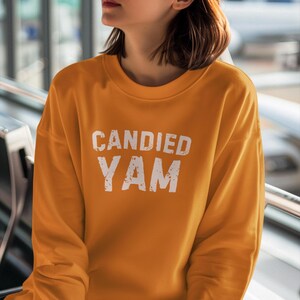 May include: Mustard yellow long-sleeve sweatshirt with the text "CANDIED YAM" in a distressed white font. The sweatshirt has a crew neck and a relaxed fit. The image is a medium shot, focusing on the upper body.