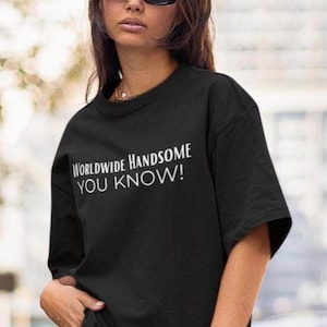 Puede incluir: Camiseta negra con el texto blanco "WORLDWIDE HANDSOME YOU KNOW!". La camiseta tiene cuello redondo y mangas cortas. La imagen es un plano medio, centrado en la camiseta y el texto.