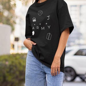 Puede incluir: Camiseta negra con elementos gr&aacute;ficos blancos: una corona, una cara sonriente, la letra "J", la palabra "ARMY", un coraz&oacute;n y un libro. La camiseta se combina con vaqueros azules.