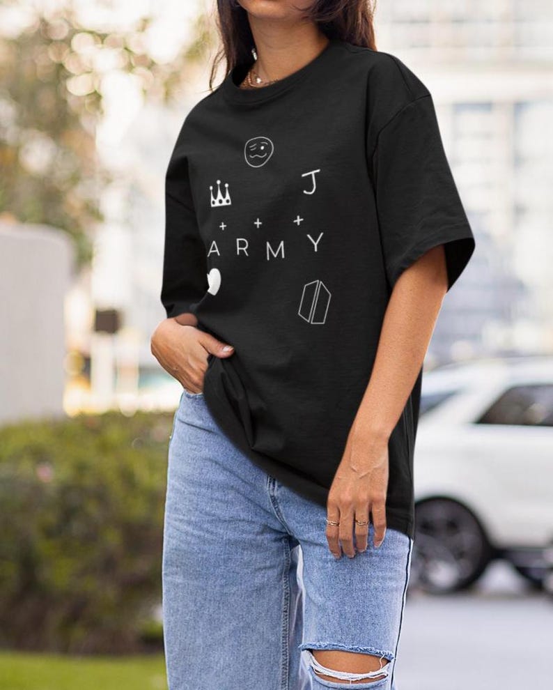 Puede incluir: Camiseta negra con elementos gr&aacute;ficos blancos: una corona, una cara sonriente, la letra "J", la palabra "ARMY", un coraz&oacute;n y un libro. La camiseta se combina con vaqueros azules.