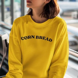 May include: A bright yellow sweatshirt with the words "CORN BREAD" printed in black across the chest. The long-sleeved top has a crew neck and a relaxed fit. The sweatshirt is made of a soft material.