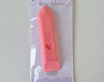 Pode incluir: Dobradeira de osso de plástico rosa para encadernação. A ferramenta tem um recorte em forma de coração e o texto "Bookbinding Tools Bone Folder" está impresso na embalagem.
