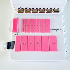 Pode incluir: Uma perfuradora branca com uma série de guias retangulares cor-de-rosa rotuladas com números de #00 a #9. O dispositivo inclui uma fila superior de seis cabeças de perfuração metálicas e uma escala de medição de 0 a 0,75 cm para alinhamento.