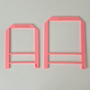 May include: Two pink plastic frames with measurements printed on them. The smaller frame measures 100 x 100 mm and the larger frame measures 130 x 130 mm.