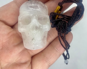 May include: A white crystal skull pendant on a black and gold cord necklace. The skull is carved with detailed features and has a smooth, polished finish.