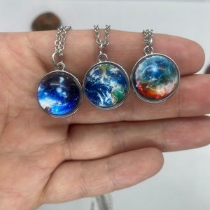 May include: Three silver necklaces with round glass pendants featuring different space-themed images. One pendant shows a blue and white image of Earth, another shows a nebula with red, orange, and blue colors, and the third shows a galaxy with a blue and white background.