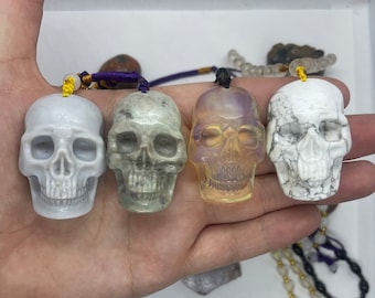 May include: Four carved skull pendants made of different types of stone. The skulls are white, green, clear, and white with black veins. Each skull has a purple cord attached to it.